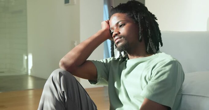African male sitting on wood floor beside white couch tilting head, reflecting as sunlight shifting