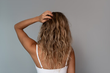 Obraz premium Back view of young blonde woman touching long wavy hair styled with curly method. Natural porous texture, voluminous bleached hairstyle and healthy hair care concept on gray background