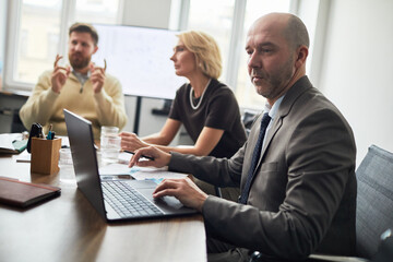 Fototapeta premium Adult businessman working on laptop during team meeting while colleagues discussing strategy in office. Suitable for corporate planning, leadership, finance marketing