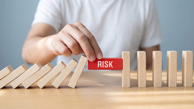 A person holding a red block labeled risk in their hand above a row of falling wooden blocks on a table