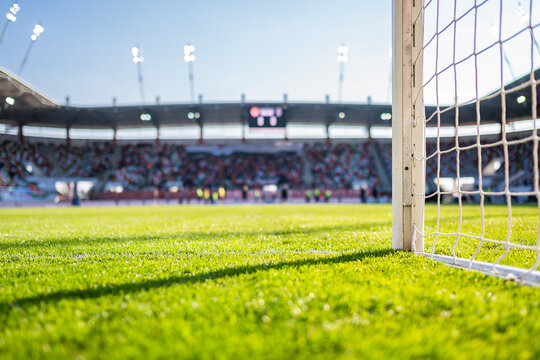 Perspective view from the goal post and net looking across a professional soccer field toward crowded stadium stands under a clear sky.