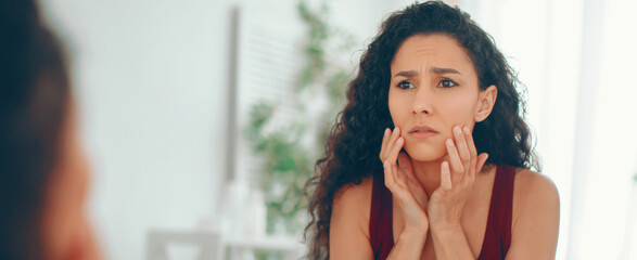 A woman stands in front of a mirror with a worried expression. She touches her cheeks with her hands. The setting appears bright and indoor. She seems to be thinking about her skin. © Prostock-studio