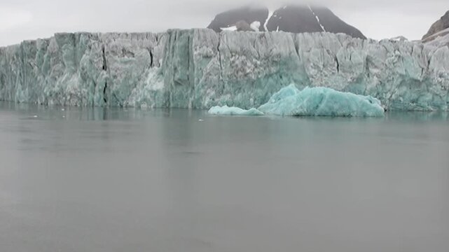Witness sublime beauty of ancient Arctic glacier under somber sky. Radiant turquoise ice formation drifts serenely upon cool, grey waters, reflecting immense scale of this polar wilderness.