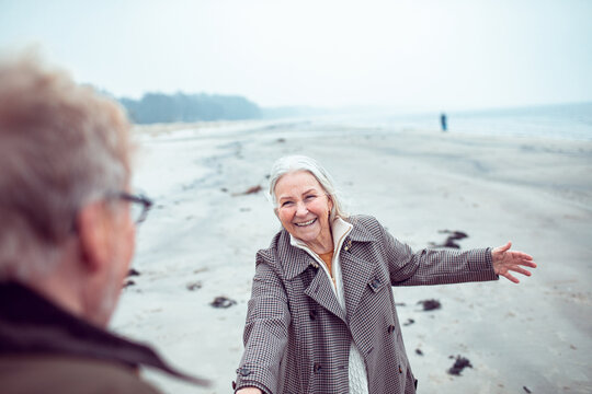 Smiling senior woman holding partner's hand on misty beach
