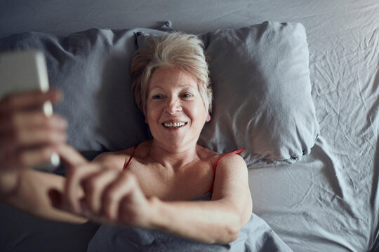 Smiling senior woman taking smartphone selfie in bed at home