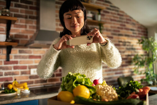 Woman photographing fresh vegetables with smartphone in home kitchen