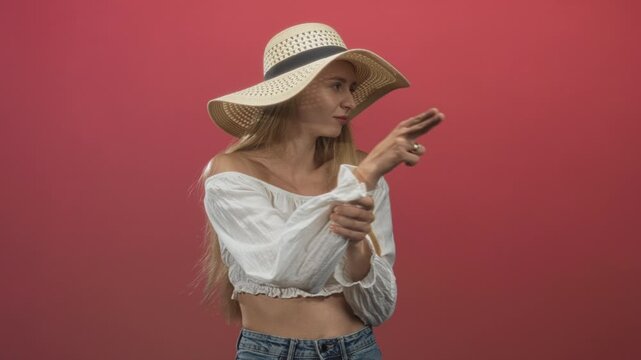 Young woman wearing straw summer hat and white off shoulder crop top, pointing finger gun while holding her wrist and showing bare midriff in studio; playful moment.