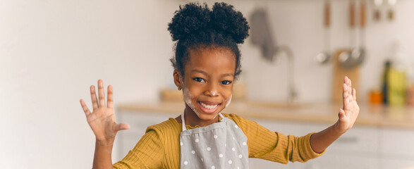 A young girl stands in a kitchen with flour on her hands. She smiles widely as she enjoys baking cookies. The kitchen has light colors and baking tools in the background. © Prostock-studio