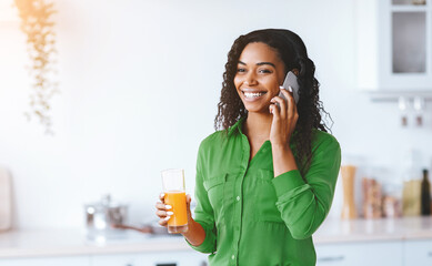 A woman stands in a kitchen while talking on her phone. She holds a glass of orange juice in her hand. The kitchen has a bright and modern look. It is daytime. © Prostock-studio