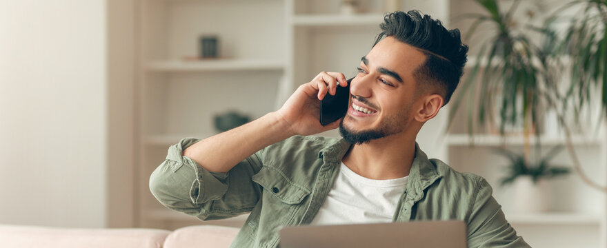 Man is sitting on a couch in a bright room. He is smiling and talking on a phone. A laptop is beside him. There are plants and shelves in the background.