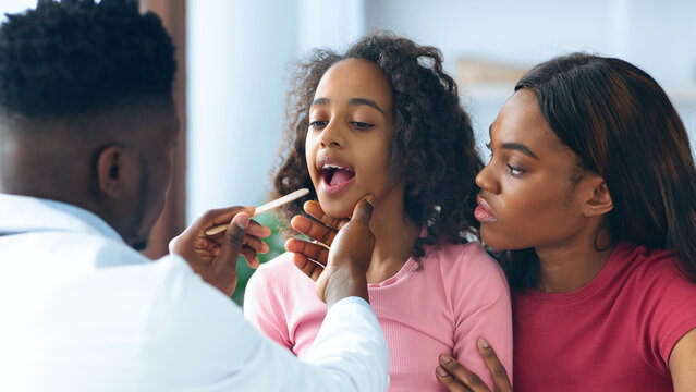 A doctor checks the throat of a young girl while her mother looks on with concern. The setting is a health clinic with a bright atmosphere during daytime.