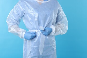 Fototapeta premium Medical worker tying disposable isolation gown on light blue background, closeup