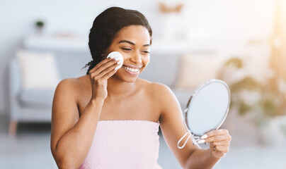A woman is using a cotton pad to apply skincare on her face while looking in a small mirror. She smiles and sits in a well-lit room with soft furniture and natural light. © Prostock-studio