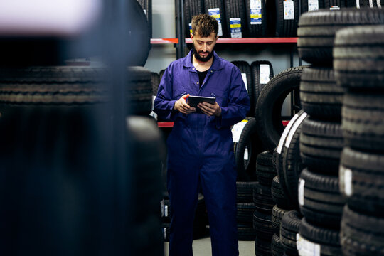 Technician in blue overalls managing tire stock and sales in a warehouse, utilizing modern technology for business operations