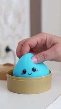 hand holding blue kawaii antistress squishy dumpling toy with cute smiley face above open plastic steamer box on white table vertical