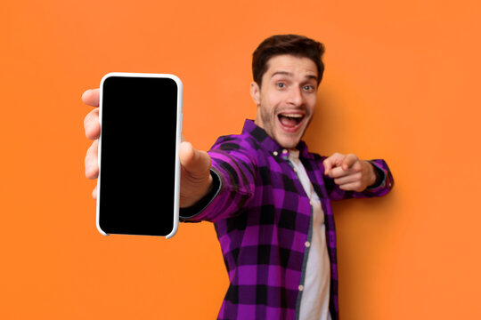 A young man stands in front of an orange wall. He smiles widely while holding a phone out. His other hand points in a friendly manner. The setting looks vibrant and cheerful.