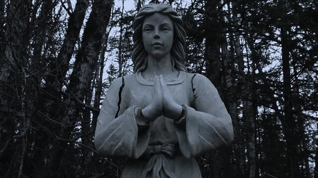 Weathered angel statue with praying hands standing in a dark forest at dusk with eerie atmospheric lighting.