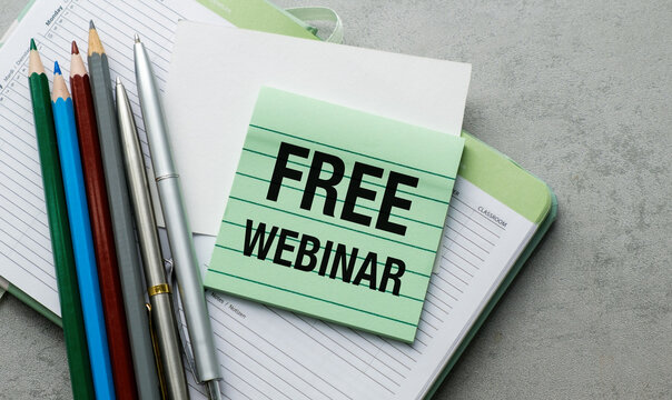 Green sticky note reads FREE WEBINAR on lined notebook. Pens and pencils rest beside the note.