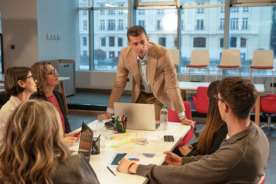 A manager stands over the table while addressing the team during a tense office meeting. The scene suggests a serious update, workplace pressure, and concerned business discussion.