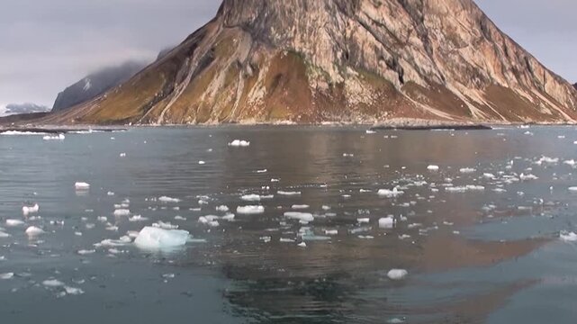 A breathtaking voyage across the Arctic Ocean. Massive ice chunks drift past towering, rugged mountains under a grey sky. An extreme travel adventure in a pristine, frozen wilderness.