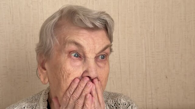 An Elderly Grandmother Covers Her Mouth With Her Hand In Shock, Sits On The Sofa In A Cozy Living Room, Pressing Her Hands To Her Lips, Wide-Eyed, Knitted Sweater. Close-up