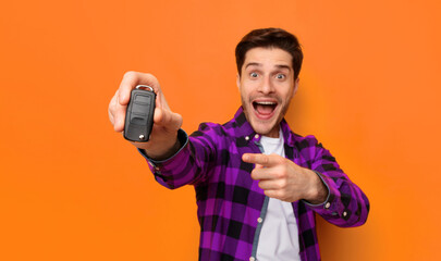 A man is smiling and pointing at car keys in his hand. He stands in front of an orange wall. His enthusiasm is clear as he expresses joy and excitement. © Prostock-studio