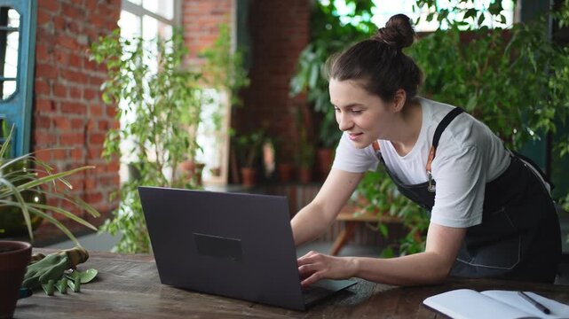 Female florist gardener working in botanical store greenhouse using laptop order plants communicating on Internet with customer. Small business owner working at flower shop. Running of own business