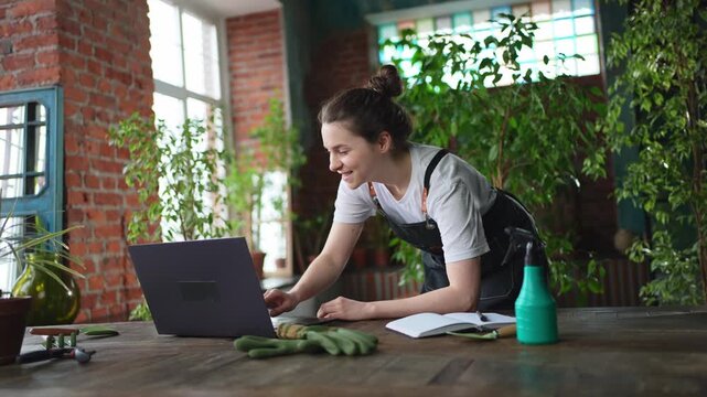 Female florist gardener working in botanical store greenhouse using laptop order plants communicating on Internet with customer. Small business owner working at flower shop. Running of own business