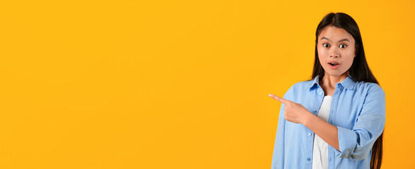 A young woman stands against a bright yellow background. She has long hair and wears a light blue shirt. She looks surprised while pointing to her right with an open expression. © Prostock-studio