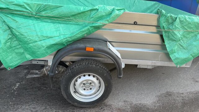 Small car trailer covered green tarp, parked roadside on asphalt near dealership, visible wheel, fender and hitch, light rust