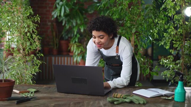 African woman florist gardener working in botanical store using laptop order plants communicating on Internet with customer. Female small business owner working at flower shop. Running of own business
