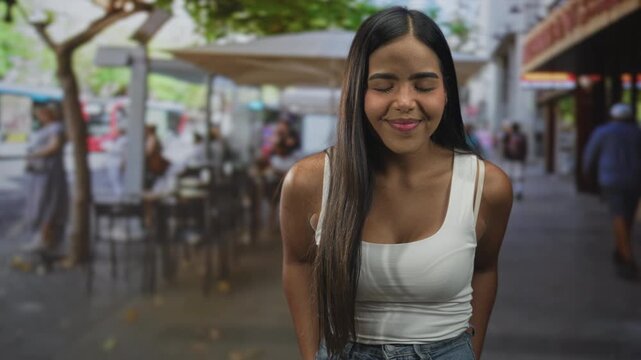 Hispanic woman in white tank top with eyes closed smiling and leaning forward at street cafe, showing bare shoulder and long hair; contentment.
