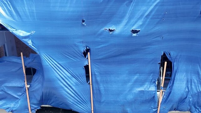 Tattered blue tarp flapping in the wind against a building facade. A damaged blue tarp is exposed to the elements, billowing and tearing in the wind, revealing glimpses of a building behind it.