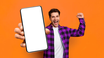 A man stands with an excited expression, holding a smartphone up with one hand. He is wearing a checkered shirt and seems to be celebrating. The background is orange. © Prostock-studio