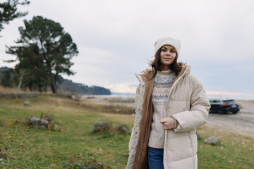Winter fashion in an outdoor setting with a woman in a beige puffer coat and knitted sweater, standing by a coastal landscape. Calm, natural light, and relaxed mood highlight warmth and casual style. © SHOTPRIME STUDIO
