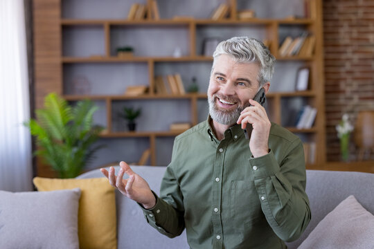 Happy mature man using a smartphone for a conversation, expressing joy and confidence while sitting comfortably at home and engaging in modern communication