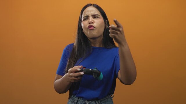 Young woman gamer holding a wireless gamepad points finger forward with a scowl and animated gesture in a studio orange backdrop during intense play; frustration.