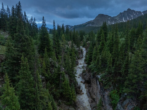 Aerial view of a rushing mountain river flowing through a rocky canyon surrounded by dense evergreen forests and rugged peaks under a moody sky CO, United States.