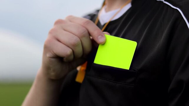 Soccer referee taking a yellow card from pocket on the field, preparing to penalize a player. Concept of rules enforcement, discipline, officiating and fair play in football.