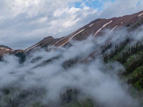 Aerial view of rugged mountain peaks with snow patches and thick fog rolling over a dense evergreen forest CO, United States.