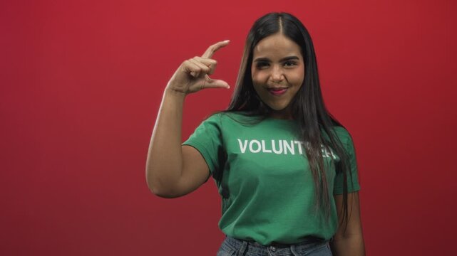 Hispanic woman wearing green volunteer shirt forms c with fingers in studio; playful confidence service spirit.