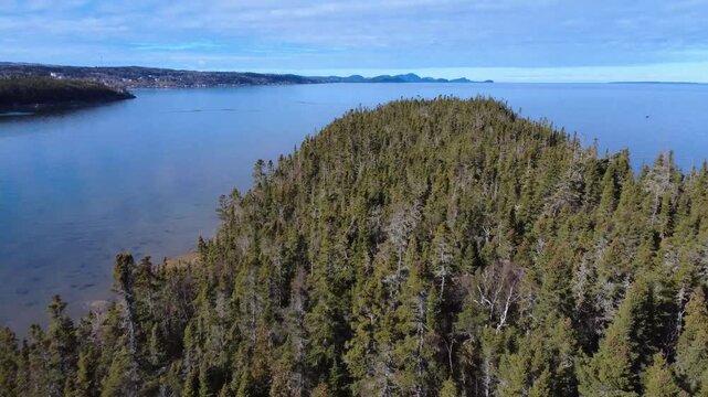 Aerial footage over dense conifer forest on Ilet Canuel facing Bic National Park peaks, with smooth St. Lawrence River waters mirroring the cloudy sky along Rimouski&rsquo;s shore.