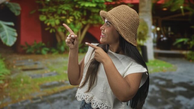 Young hispanic woman points both index fingers to upper left while smiling beside building, wearing straw hat and white lace top; joyful curiosity.
