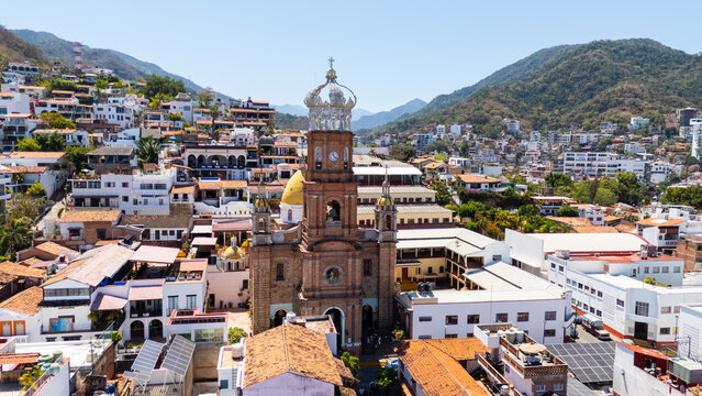 Aerial view of the Parish of Our Lady of Guadalupe with its iconic crown tower surrounded by terracotta rooftops and lush green hills under a clear blue sky in Puerto Vallarta, Jalisco, Mexico.