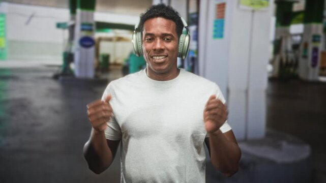 Black man wearing headphones dancing and smiling with hands raised at a street gas station; joy music energy.