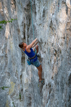 Young woman climbs a steep limestone route while secured on rope and equipped with climbing gear. Her focused expression and body position show challenge and excitement of outdoor sport climbing.