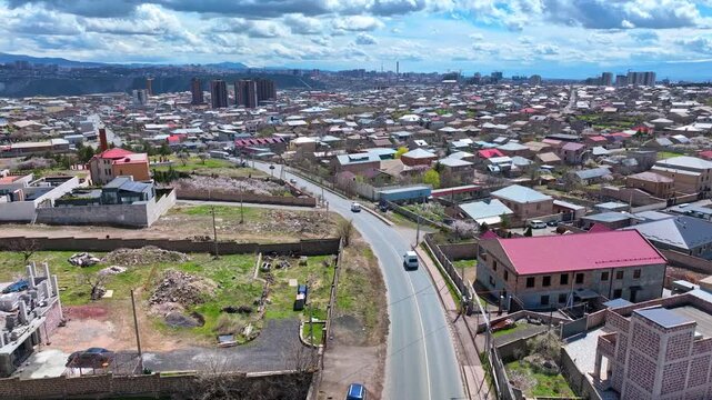 Aerial view of a cityscape with roads and houses. Clouds drift over distant mountains. Vehicles move along winding streets. Urban development spreads toward hills in background