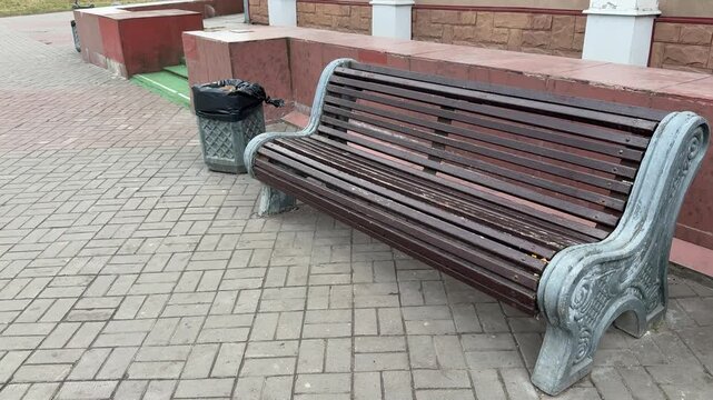 Empty park bench next to trash bin with black bag on gray paving stones during day, decorative cast iron bench ends, red low wall