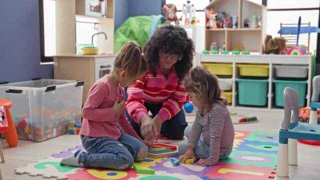 Woman helps two girls place wooden shapes on foam mat in preschool classroom; nurturing learning play.