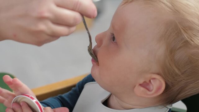 White baby being fed with spoon, closeup profile shot, caregiver hand offering puree, bib smeared, curious expression, family bonding, gentle nurturing, everyday mealtime routine